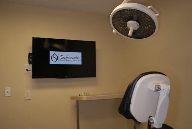Modern dental operatory with a patient chair, overhead medical lighting, and wall-mounted monitors.