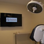 Modern dental operatory with a patient chair, overhead medical lighting, and wall-mounted monitors.